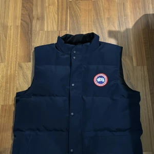 Svart dunväst från Canada Goose - Säljer en snygg och varm svart dunväst från Canada Goose. Västen har en quiltad design med knappar framtill och en hög krage för extra värme. Perfekt för kyliga höst- och vinterdagar. Den är i storlek M och i mycket bra skick.