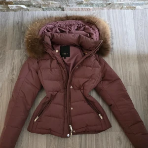 Brun dunjacka från Saki - Säljer en snygg dustpink dunjacka från Saki med en fluffig päls på huvan. Jackan har en dragkedja och knappar framtill, vilket gör den perfekt för kalla vinterdagar. Den är figursydd för en smickrande passform och har långa ärmar. Perfekt för både vardag och fest!