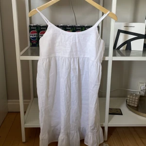 Vit linneklänning från Gina Tricot - Säljer en superfin vit linneklänning från Gina Tricot. Den är ärmlös med smala axelband och har en luftig passform. Perfekt för sommardagar eller en avslappnad fest. Klänningen har en söt volangdetalj längst ner som ger den lite extra stil. Materialet är linne, vilket gör den sval och bekväm att bära.