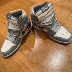 Grå och vita sneakers från Nike x Dior - Exklusiva sneakers i samarbete mellan Nike och Dior. Skorna har en stilren design i grått och vitt med Dior-logotypen på swooshen. Högt skaft med snörning och perforerad tå för extra ventilation. Perfekta för den som vill ha något unikt och trendigt men även lite lyx. 