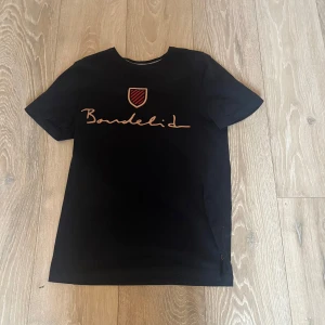 Svart t-shirt från Bondelid - Snygg svart t-shirt från Bondelid med ett stilrent tryck i orange på bröstet. Perfekt för en casual look. Tillverkad i mjukt material för extra komfort.