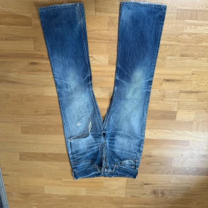 Blå jeans från Levi's - Säljer ett par klassiska blå jeans från Levi's med slitna detaljer och hål. De har en straight passform och är tillverkade i denim. Perfekta för en avslappnad stil. Vintage Levis 517