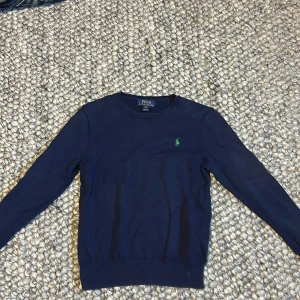 Mörkblå tröja från Ralph Lauren - Säljer en stilren mörkblå tröja från Ralph Lauren med det klassiska gröna logotypbroderiet på bröstet. Tröjan har långa ärmar och en rund halsringning. Perfekt för en avslappnad och snygg look. Storlek M i barn storlek vilket passar 12 år 