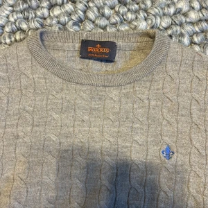 Kabelstickad tröja från Morris - Säljer en stilren grå kabelstickad tröja från Morris. Tröjan har en rund halsringning och ett diskret broderat emblem på bröstet. 100 merinoull