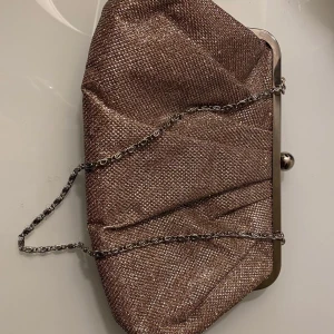 Brun glittrig kuvertväska från Accessorize - Snygg brun glittrig kuvertväska från Accessorize med en elegant kedja som axelrem. Väskan har en metallram och ett knäppe upptill. Insidan är fodrad med ett matchande brunt tyg och har en liten ficka för extra förvaring. Perfekt för att ge en touch av glamour till din outfit!❣️💫