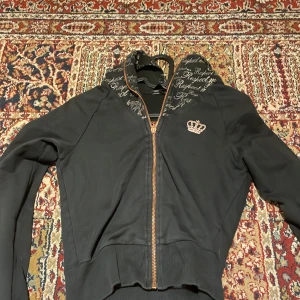 Svart huvtröja från Adidas - Säljer en svart huvtröja från Adidas med en snygg krona på bröstet och textmönster på huvan. Tröjan har dragkedja och långa ärmar, perfekt för en casual look. Huvan ger extra stil och värme.