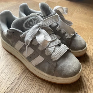 Grå Adidas campus 00s - Snygga grå sneakers från Adidas i mocka med vita detaljer och snörning. De har den klassiska tre-randiga designen på sidorna och en bekväm sula. Perfekta för en avslappnad stil.