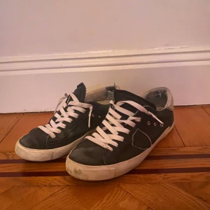 Phillipe model - Säljer ett par svarta sneakers med vita snören och sulor. Skorna har en klassisk design med lågt skaft och är perfekta för en avslappnad stil. De har en enkel och stilren look som passar till det mesta. Ingen låda