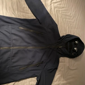 Svart jacka med huva och glasögon - Säljer en marinblå jacka från C.P. Company med inbyggda glasögon i huvan. Jackan har dragkedja framtill och flera praktiska fickor med dragkedjor. Perfekt för en unik och stilren look. 