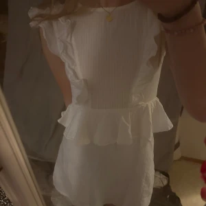 Vit klänning med volangdetaljer - Säljer en söt vit klänning med volangdetaljer vid axlarna och midjan. Klänningen har en kort längd och är perfekt för sommarens varma dagar. Den är ärmlös och har en fin passform som framhäver figuren. Perfekt för studenten eller sommaren! 🌸