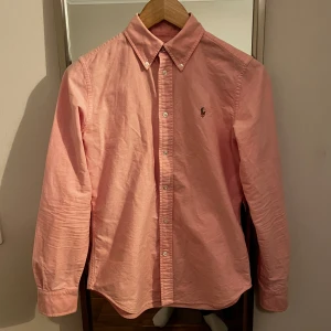 Rosa skjorta från Ralph Lauren - Säljer en stilren rosa skjorta från Ralph Lauren i super slim fit. Skjortan har långa ärmar och knappar framtill. Den är broderad med den klassiska loggan på bröstet. Perfekt för en snygg och avslappnad look. Riktigt bra skick sitter som storlek S
