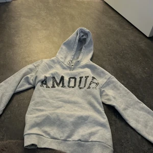 Grå hoodie från Gina Tricot med tryck - Säljer en grå hoodie från Gina Tricot med texten 'AMOUR' tryckt på framsidan och en stjärna på baksidan. Tröjan har en klassisk huva med dragsko och långa ärmar. Perfekt för en avslappnad stil.