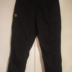 Svarta byxor från Fjällräven - Säljer ett par svarta byxor från Fjällräven med praktiska fickor och dragkedja. Perfekta för utomhusaktiviteter med sin robusta design och bekväma passform. Helt oanvända pga fel storlek. 