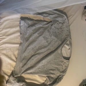 Grå tröja med pärlor från Lindex - Säljer en stilren grå tröja från Lindex med pärldetaljer på axlarna. Tröjan har långa ärmar och är perfekt för en avslappnad look. Tillverkad i ett mjukt material som känns skönt mot huden.