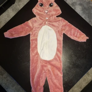 Rosa kanindräkt med huva - Mysig rosa kanindräkt med huva och vita detaljer. Dräkten har en dragkedja framtill och en söt vit svans baktill. Perfekt för en rolig och bekväm utklädnad.