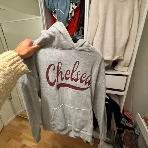 Grå hoodie med Chelsea-tryck - Säljer en grå hoodie med stort Chelsea-tryck i vinrött på framsidan. Tröjan har en klassisk känguruficka och en bekväm huva. Perfekt för en avslappnad stil. Hör av dig vid intresse!