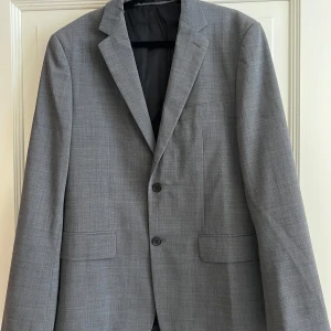 Grå kavaj från Riley - Snygg grå kavaj från Riley i en stilren design. Kavajen har en skräddarsydd passform med två knappar framtill och klassiska slag. Tillverkad i en blandning av ull och polyester för en bekväm känsla.