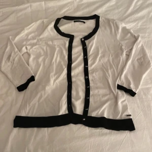 Elegant kofta från MOHITO - Säljer en stilren vit kofta från Mohito med svarta detaljer. Koftan har långa ärmar och knappar framtill. Perfekt för en elegant look.