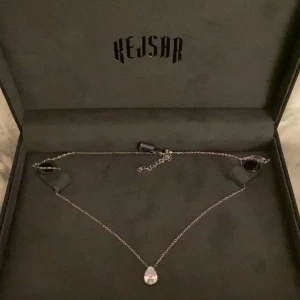 Silverhalsband med droppformad hängsmycke från Kejsar - Elegant silverhalsband från Kejsar med ett glittrande droppformat hängsmycke. Perfekt för att lägga till en touch av glamour till din look. Levereras i en stilren svart ask med Kejsar-logotypen i guld.