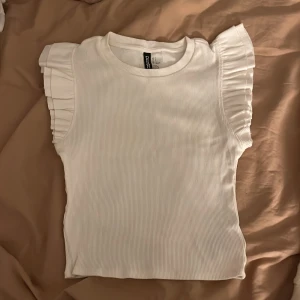 Vit ribbad topp från H&M - Säljer en söt vit ribbad topp från H&M med volangdetaljer på ärmarna. Säljer pga inte kommer till användning