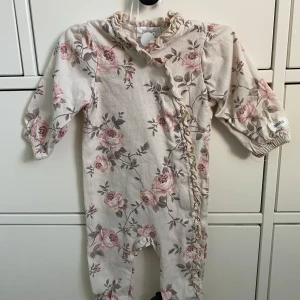 Blommig onesie från ytan - Söt onesie från Newbie i beige med rosa blommönster. Den har långa ärmar och volangdetaljer längs knäppningen. Perfekt för mysiga dagar.