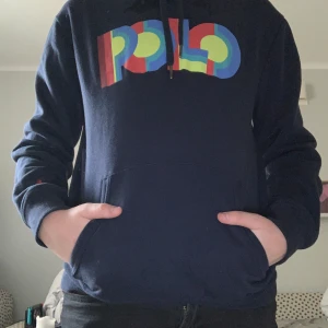 Mörkblå hoodie från Polo Ralph Lauren - Säljer en snygg mörkblå hoodie från Polo Ralph Lauren med färgglatt POLO-tryck på bröstet. Den har en klassisk känguruficka och en liten röd logga på ärmen. Perfekt för en avslappnad stil.