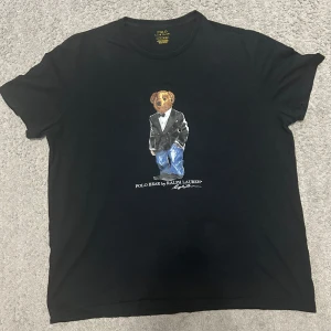 Svart t-shirt med Polo Bear från Ralph Lauren - Snygg svart t-shirt från Ralph Lauren med det ikoniska Polo Bear-motivet på framsidan. T-shirten har en klassisk passform och är tillverkad i mjuk bomull för en bekväm känsla. Perfekt för en stilren och avslappnad look.