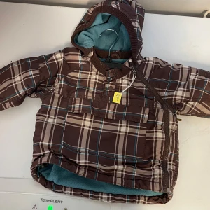 Brun rutig vinterjacka med huva - Säljer en brun rutig vinterjacka till baby 1-2 år med huva och dragkedja. Stor i storleken. Jackan har en mysig fleecefoder i turkos och en praktisk ficka framtill. Perfekt för kalla dagar!