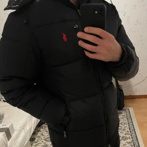 Svart dunjacka från Ralph Lauren - Säljer en svart dunjacka från Ralph Lauren med röd broderad logga på bröstet. Jackan har en huva och dragkedja framtill. Perfekt för kyliga dagar med sin pufferdesign som håller värmen. Knappt använd S/M