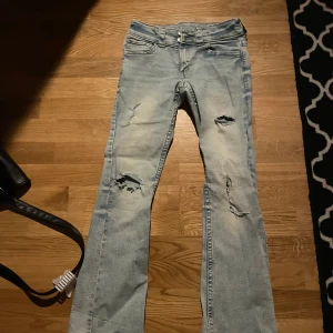 Ljusa jeans med slitningar från H&M - Snygga ljusa jeans från H&M med slitningar på benen för en trendig look. De har en bootcut-stil och är lågmidjade med justerbar midja. Perfekta för en avslappnad stil.