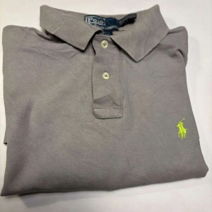 Grå pikétröja från Polo Ralph Lauren - Säljer en klassisk grå pikétröja från Polo Ralph Lauren med korta ärmar och en broderad logotyp i gult på bröstet. Perfekt för en stilren och avslappnad look. Tröjan har en traditionell krage och knappar framtill.