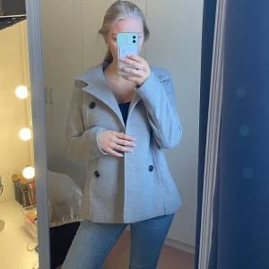 Grå kappa med knappar - Säljer en stilren grå kappa med dubbelknäppning och stora svarta knappar. Jackan har en klassisk krage och långa ärmar, perfekt för kyligare dagar. Den är i en snygg och tidlös design som passar till många olika outfits. Den är i storlek M och passar mig som bär S. 