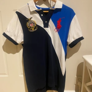 Flerfärgad pikétröja från Ralph Lauren - Snygg flerfärgad pikétröja från Ralph Lauren med klassisk krage och knappar. Tröjan har ett stort rött broderat logotypmotiv på bröstet och en emblem på ena sidan. Perfekt för en stilren look.