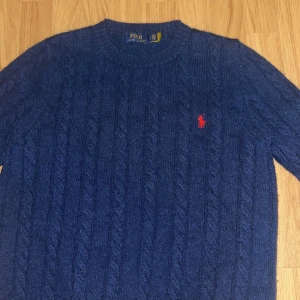 Blå stickad tröja från Polo Ralph Lauren - Säljer en snygg blå stickad tröja från Polo Ralph Lauren med det klassiska röda logotypen på bröstet. Tröjan har en rund hals och långa ärmar, perfekt för kyligare dagar. Passar både till jeans och chinos för en stilren look.