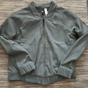 Grå träningsjacka från Adidas - Säljer en grå ä/grön träningsjacka från Adidas med dragkedja framtill. Jackan har långa ärmar och en diskret prickig textur. Perfekt för träning eller en avslappnad stil. Adidas-loggan finns på ärmen.