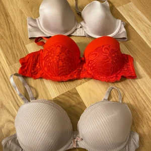 Tre olika BH:ar i olika färger och stilar strl 75B - Säljer tre BH:ar i olika stilar och färger. En beige med slät yta, en röd med spetsdetaljer och en beige med diskreta ränder och liten rosett framtill. Alla har justerbara axelband och bygel för extra stöd.
