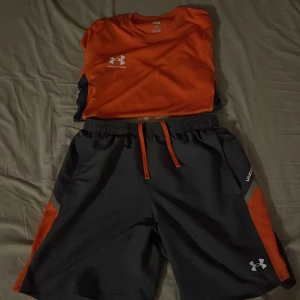 Orange tracksuit från Under Armour - Snygg orange t-shirt från Under Armour med korta ärmar och kontrasterande mörkblå sidopaneler. Perfekt för träning eller en aktiv dag. Logotypen är tryckt på bröstet.