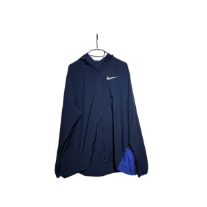 Mörkblå vindjacka från Nike - Snygg mörkblå vindjacka från Nike med huva och dragkedja. Jackan har en stor Nike-logga på bröstet och texten 'NIKE RUNNING' längs ärmen.
