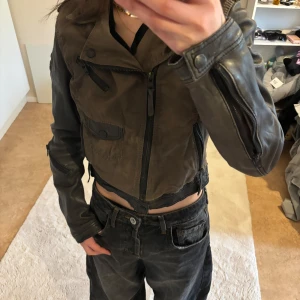 Biker jacka st: S - Säljer en cool brun och svart skinnjacka från Tigha. Jackan har en asymmetrisk dragkedja och flera fickor med dragkedjor. Den har långa ärmar och en stilren design med detaljer som knappar på axlarna.