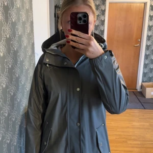 Grå regnjacka från Swedemount - Snygg grå regnjacka från Swedemount med huva och dragkedja. Jackan har knappar framtill och justerbara snören vid huvan. Perfekt för regniga dagar och håller dig torr och bekväm. Sparsamt använd, gott skick