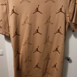 Beige t-shirt med mönster Nike Jordan - Snygg beige Nike Jordan  t-shirt med ett ikoniskt mönster över hela tyget. Perfekt för en avslappnad stil. Kortärmad och i mjukt material för extra komfort.