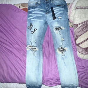 Blå jeans med slitningar från Amiri - Säljer ett par coola blå jeans från Amiri med slitningar och broderade detaljer. Jeansen har en modern look med en ljus tvätt och är perfekta för en trendig stil. De har en klassisk femficksdesign och knappgylf.