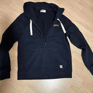 Mörkblå hoodie från Dobber - Säljer en snygg mörkblå hoodie från Dobber med dragkedja och vita snören. Den har en broderad logga på bröstet och praktiska fickor framtill. Perfekt för en avslappnad stil. Mycket gott skick, använd sparsamt, tvättar ett par ggr. Storlek M