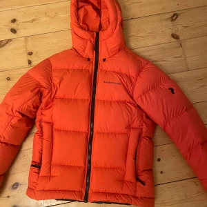 Orange dunjacka från Peak Performance - Säljer en snygg orange dunjacka från Peak Performance. Jackan har en dragkedja framtill och en praktisk huva. Perfekt för kyliga dagar med sin varma och puffiga design. Två fickor med dragkedja på sidorna för extra förvaring.