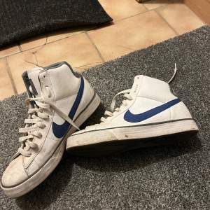 Säljer ett par vita Nike sneakers med blå swoosh och högt skaft. Skorna har snörning och en klassisk design med Nike-logga på hälen. Perfekta för en sportig look.