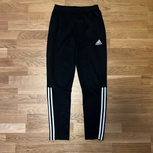 träningsbyxor Adidas - Snygga svarta träningsbyxor från Adidas med klassiska vita ränder längs sidorna. Byxorna har en elastisk midja och en smal passform som ger en sportig look. Perfekta för träning eller en avslappnad dag. Passar en person från 160 - 170cm.