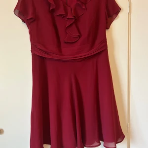 Vinröd klänning med volangdetalj - Elegant vinröd klänning med volangdetalj framtill och korta ärmar. Perfekt för speciella tillfällen med sin vackra design och flödande kjol. Dragkedja baktill för enkel påklädning.