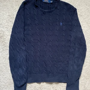 Mörkblå stickad tröja från Polo Ralph Lauren - Säljer en snygg mörkblå stickad tröja från Polo Ralph Lauren med kabelstickat mönster. Tröjan har långa ärmar och en liten broderad logga på bröstet. Perfekt för kyligare dagar!