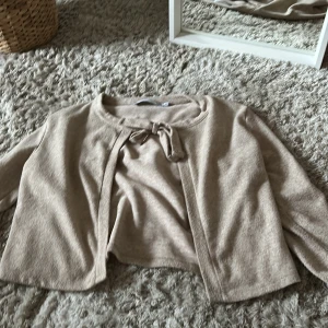 Beige kofta från 157 - Säljer en stilren beige kofta från 157 i storlek XS. Koftan har långa ärmar och en knytning framtill som ger en fin detalj. Perfekt för lager-på-lager under kyligare dagar.