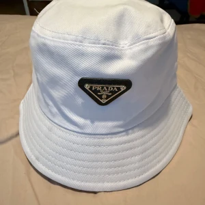 Vit hatt från Prada - Snygg vit hatt från Prada med en klassisk design. Hatten har en bred kant och ett elegant märke framtill. Perfekt för att ge en stilren touch till din outfit.